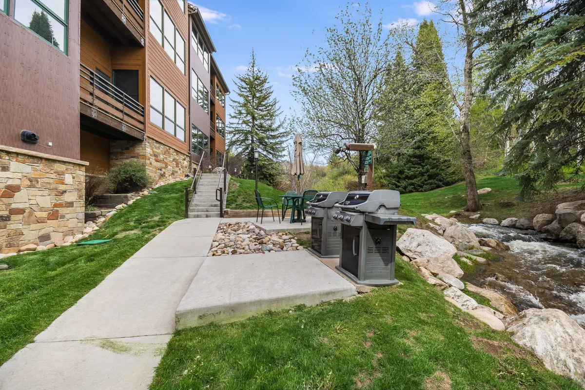 The landscaped yard features a grill, seating area, and the nearby stream, inviting residents and guests to relax, dine, or play on the grass. Children can be seen enjoying the lawn while adults gather for a barbecue, set against a backdrop of alpine greenery.