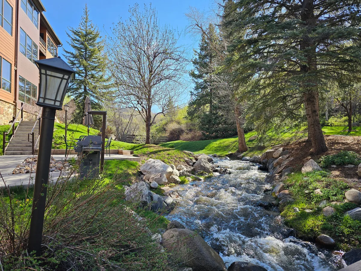 A serene creek flows beside a lush lawn and patio area, framed by landscaped trees. The tranquil setting is perfect for relaxing after skiing, enjoying a winter sunset, or sipping coffee by the water.
