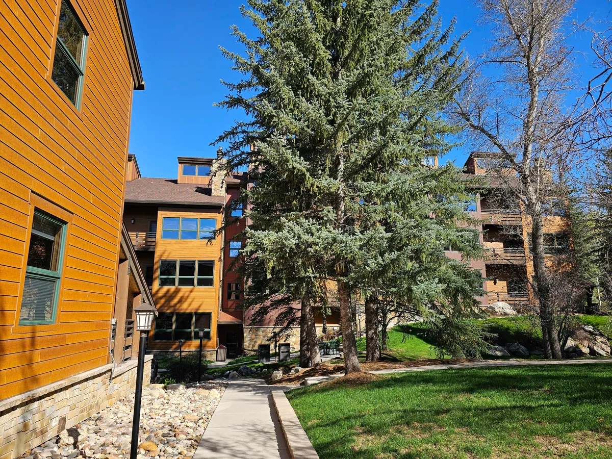 Colorful wooden buildings line a landscaped pathway, with a tall evergreen and outdoor lighting providing a cheerful, active outdoor environment. The setting is ideal for strolls, socializing, and enjoying the mountain air after a day on the slopes.