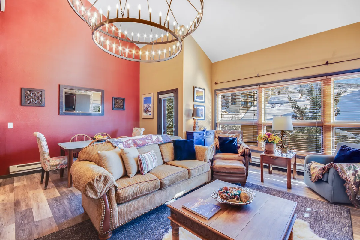 A view of an integrated living and dining area featuring a cozy sofa, a spacious dining table, and a window framing peaceful outdoor views. The space is adorned with a chandelier and offers a sense of warmth and togetherness, ideal for family meals or relaxing with a book.