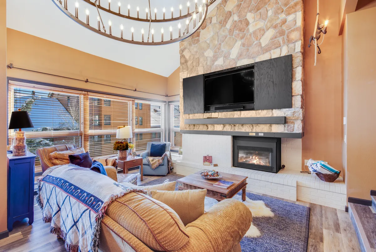 A spacious and inviting living area featuring a striking stone fireplace, a large chandelier overhead, comfortable seating, and expansive windows that fill the room with natural light and offer a glimpse of the outdoors.