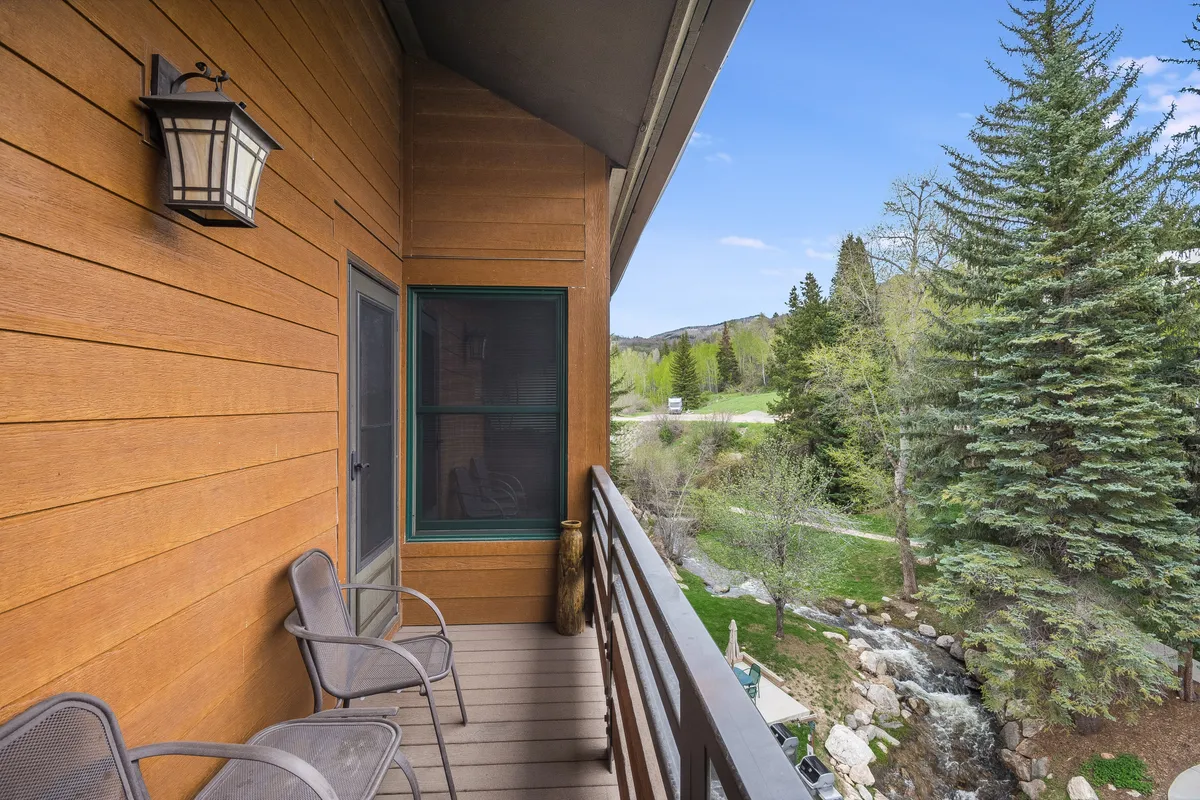 An outdoor wooden balcony overlooking lush greenery, a gentle stream, and distant mountains, creating a tranquil setting perfect for relaxation, birdwatching, or reading while embraced by nature.