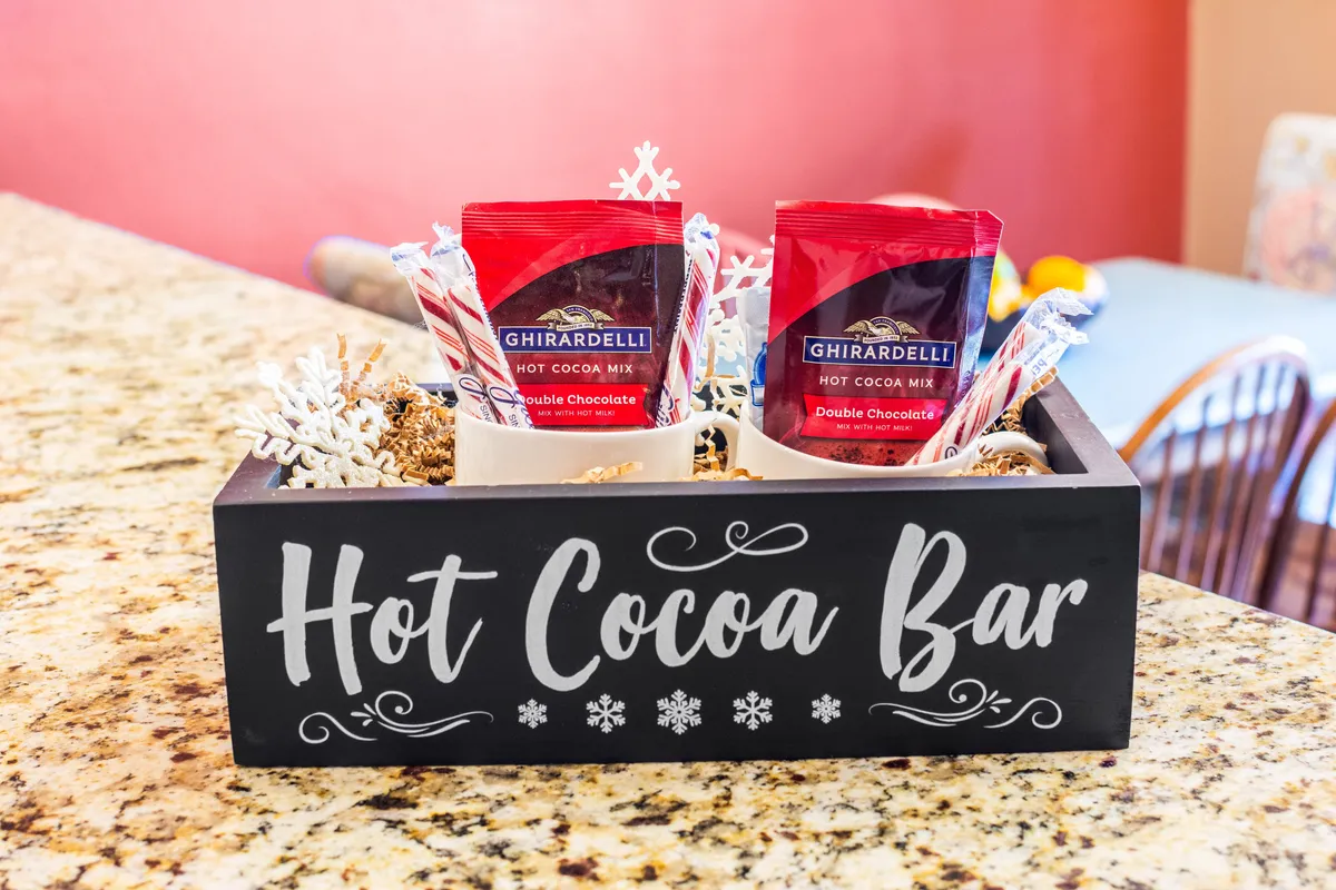 A festive wooden 'Hot Cocoa Bar' with Ghirardelli cocoa packs, spoons, and decorative snowflakes, evoking a cozy, sociable après-ski atmosphere ideal for gathering after a day on the slopes.