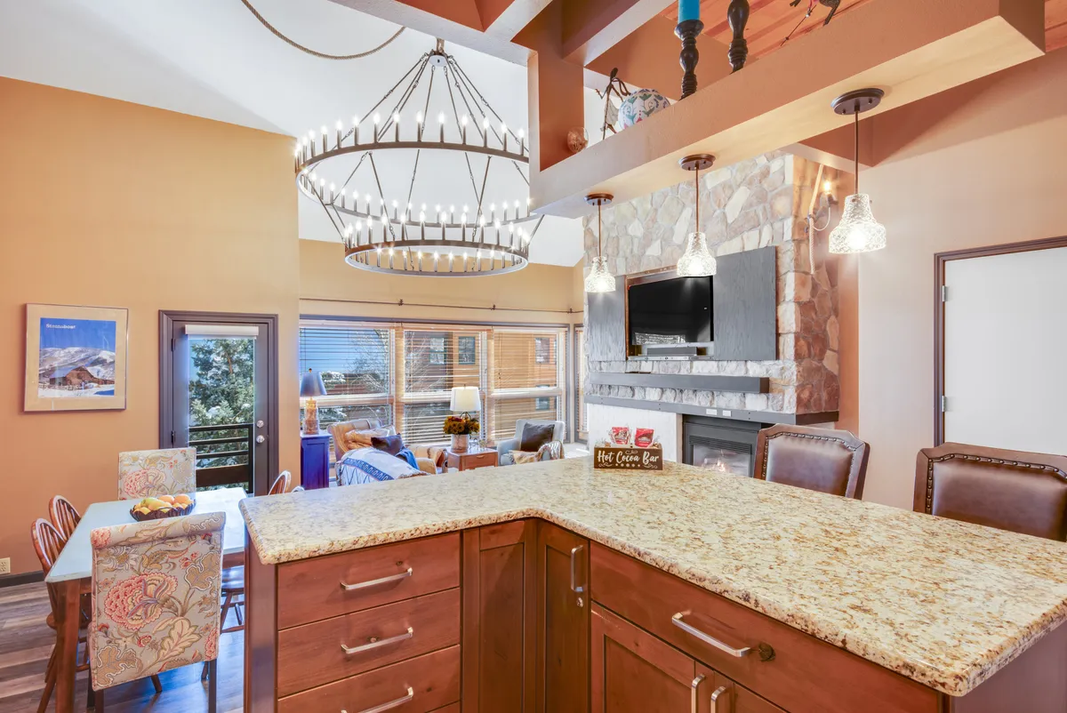 A warm, welcoming open-plan living and dining area featuring a rustic chandelier, a stone fireplace, and a large dining table set for a group. The scene captures a space perfect for family gatherings, group meals, and relaxed evenings together.