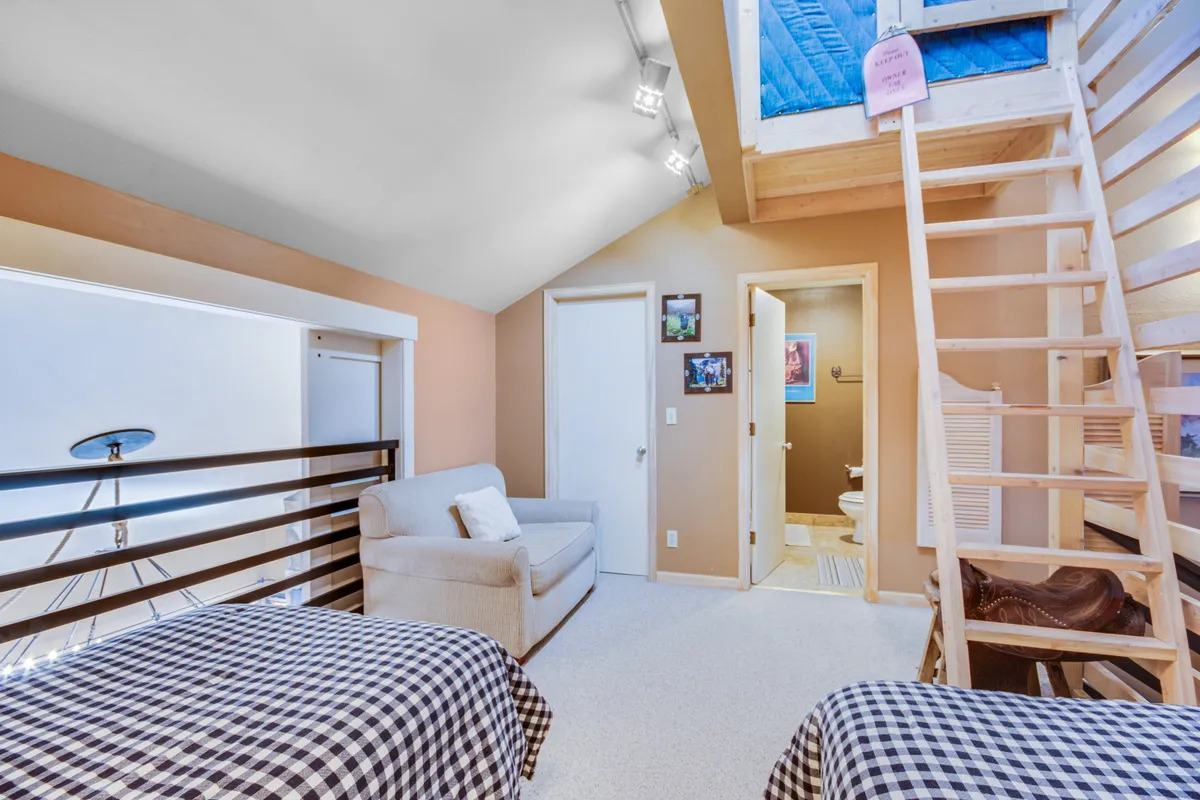 A semi-enclosed loft space featuring two twin beds with checkered bedding, a cozy armchair, and wooden ladder access. The casual atmosphere is ideal for kids or friends, emphasizing the flexible sleeping arrangements and the playful, social nature of the loft.