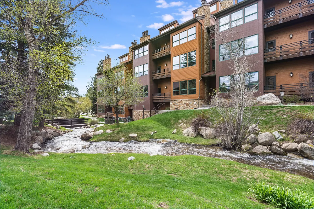This image captures the exterior of the Kutuk building with the flowing stream running alongside it, lush green grass, and balconies overlooking the peaceful landscape. The natural setting is serene, with a clear view of the water, grassy area, and the building itself nestled amid alpine scenery.