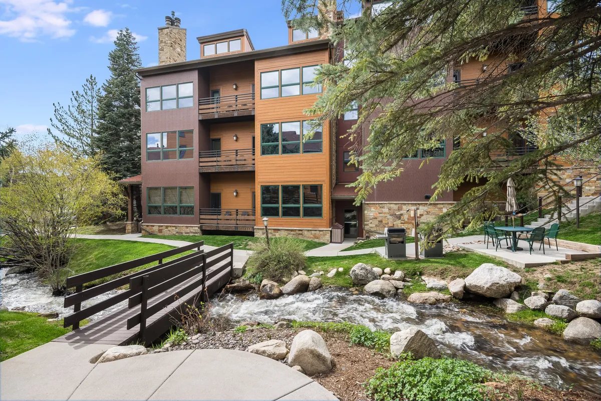 This exterior image shows a three-story building set beside a serene stream, with a wooden bridge leading to a patio area equipped with chairs. The landscaped setting is peaceful, inviting residents to relax by the water or enjoy outdoor meals.