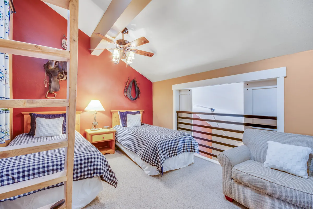 A comfortable, family-friendly loft space featuring two twin beds with checkered bedding, a cozy sofa with pillows, a wooden ladder, a table lamp, and playful wall decor. The arrangement is inviting and ideal for kids or extra guests, offering flexibility and a casual vibe.