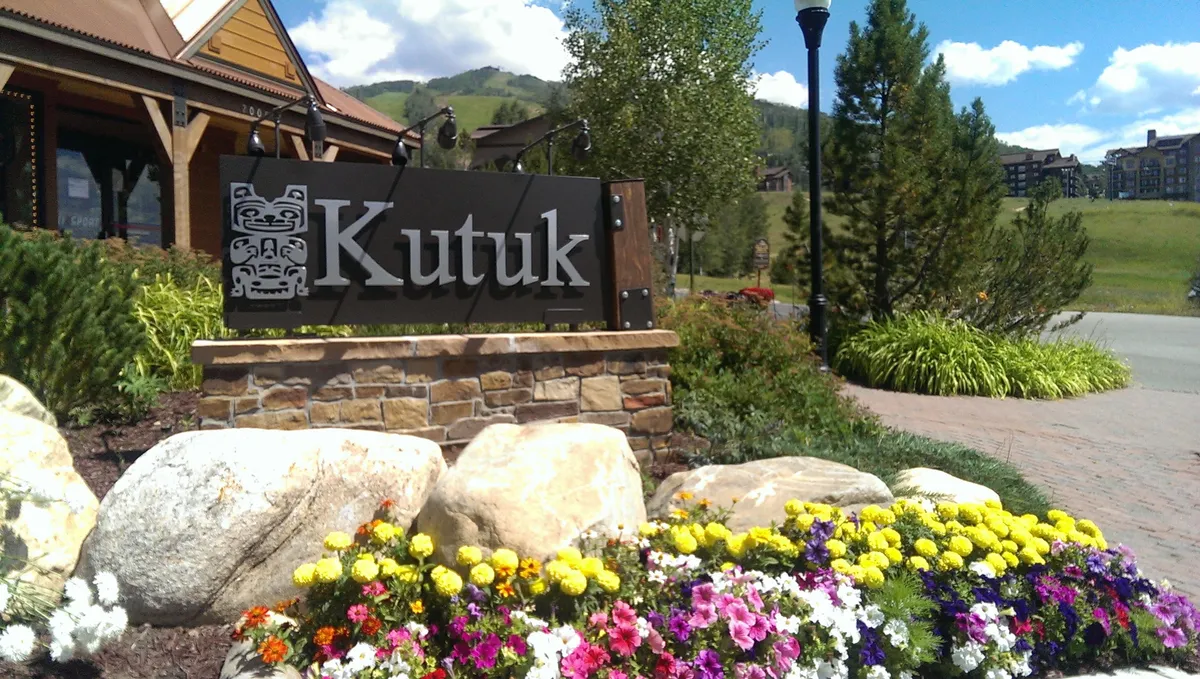 The exterior of the Kutuk property is shown with a prominent Kutuk sign, vibrant flower beds, and well-maintained landscaping. The architecture reflects a welcoming mountain lodge vibe, inviting guests to begin their adventure steps from the slopes.