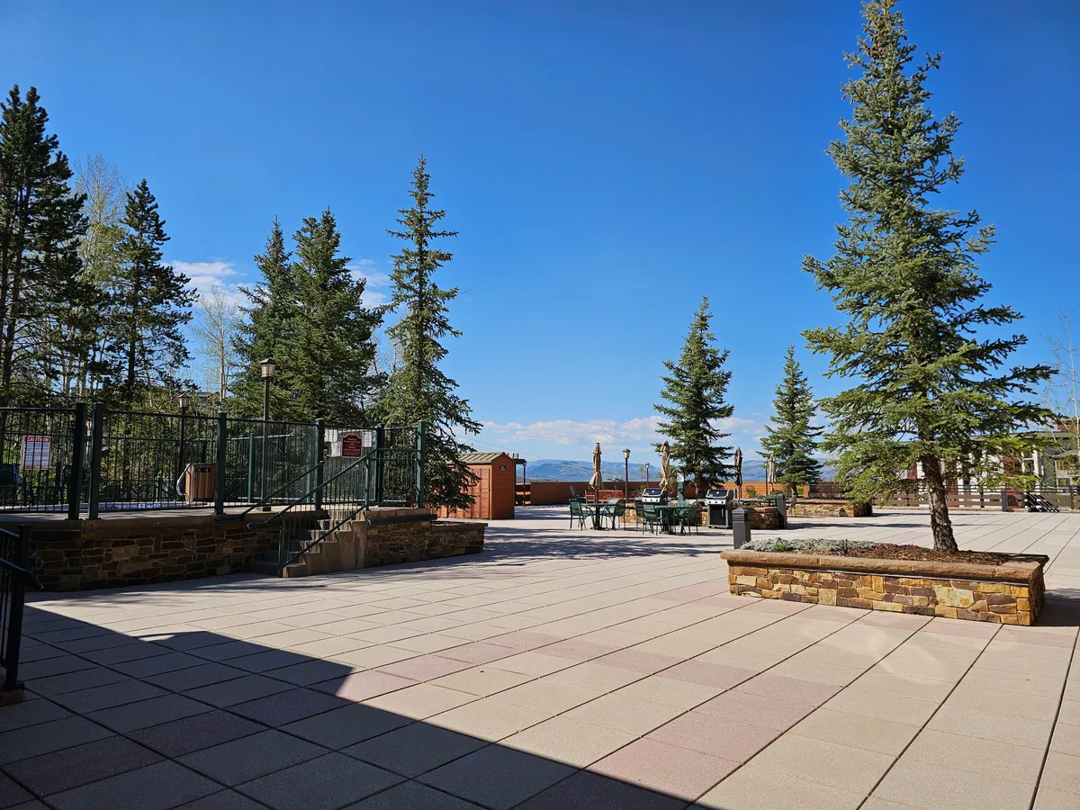 The image features a tranquil exterior community space with tall evergreen trees, an inviting outdoor seating area, stone planters, and a fenced section. The setup suggests relaxation, socializing, and a communal atmosphere perfect for gatherings or enjoying nature.