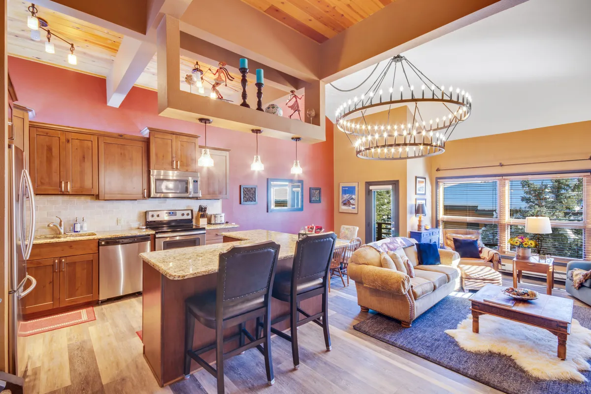 This image showcases the condo's inviting open-concept living space, featuring a stylish kitchen island under an elegant chandelier, a cozy living room seating area perfect for gatherings, and expansive windows that let in mountain light and offer views of the surrounding landscape.